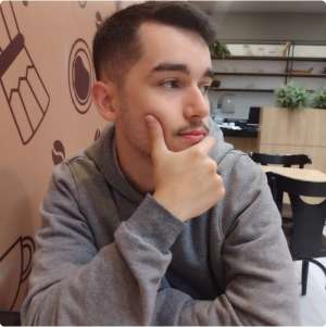 A profile picture of a young man with short hair, wearing a gray hoodie, looking to the side with his hand on his chin, sitting in a café with wall decorations featuring drawings of cups.